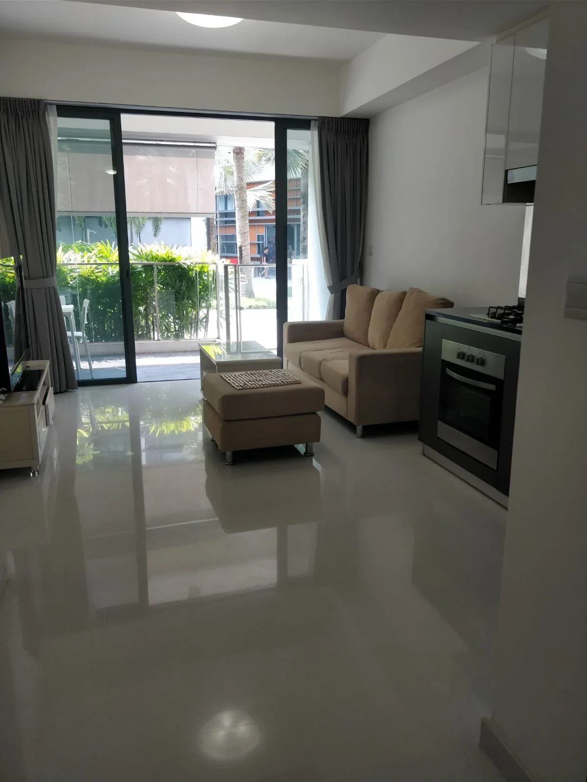 Living room with beige sofa, ottoman, glass coffee table, TV, and sliding glass door leading to a balcony with outdoor furniture and greenery.