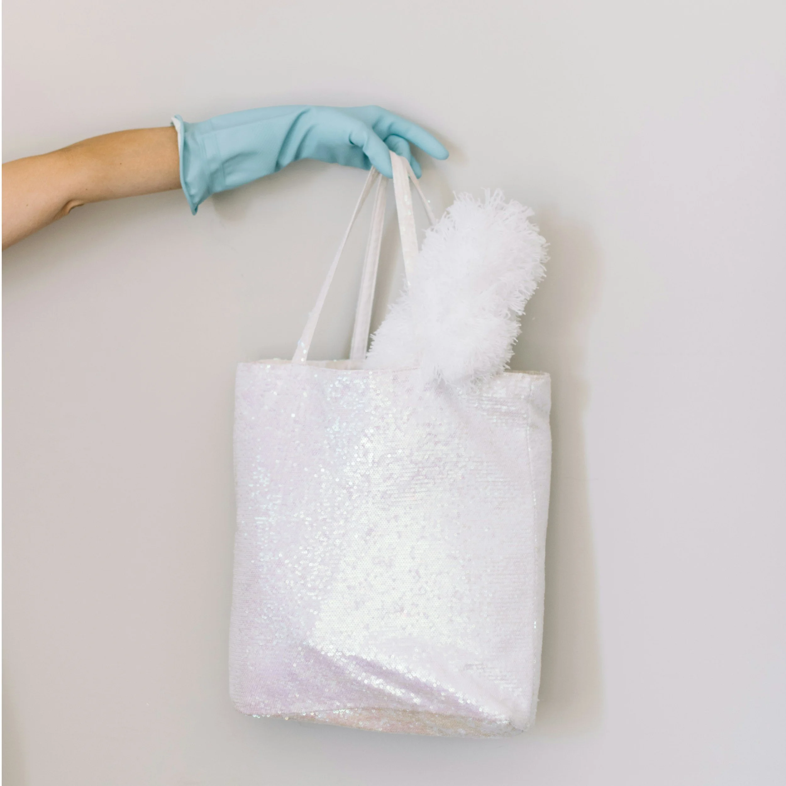 A hand wearing a blue glove holding a white, glittery tote bag on a plain white background.