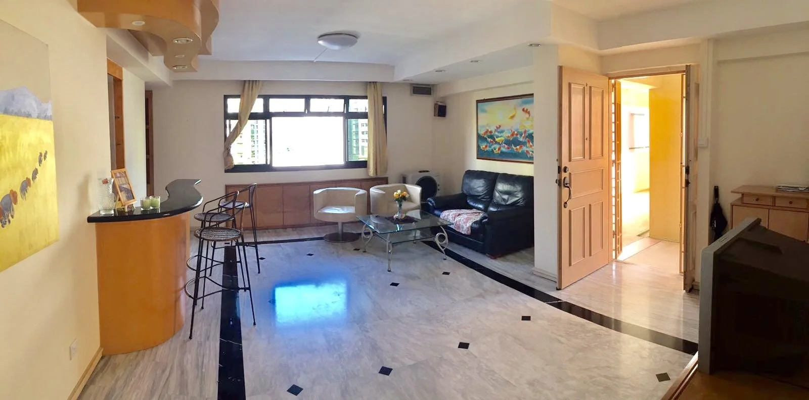 Living room with yellow walls, a black leather couch, white and beige chairs, a glass coffee table with a flower vase, large window with cream curtains, artwork on the walls, a wooden door open to a balcony, and a small bar area with bar stools.