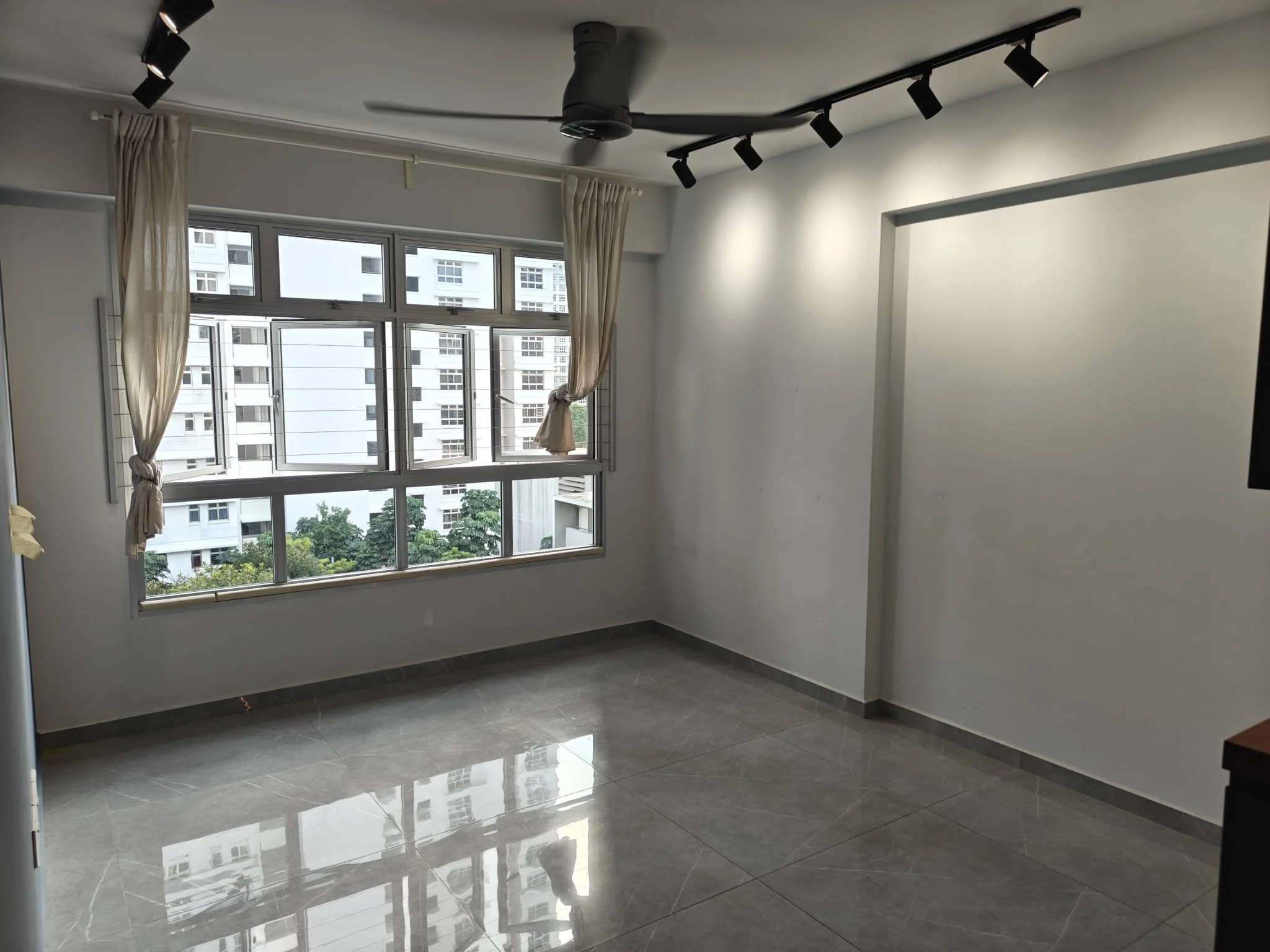 Empty room with large window, beige curtains, gray tiled floor, white walls, ceiling fan, and track lighting.