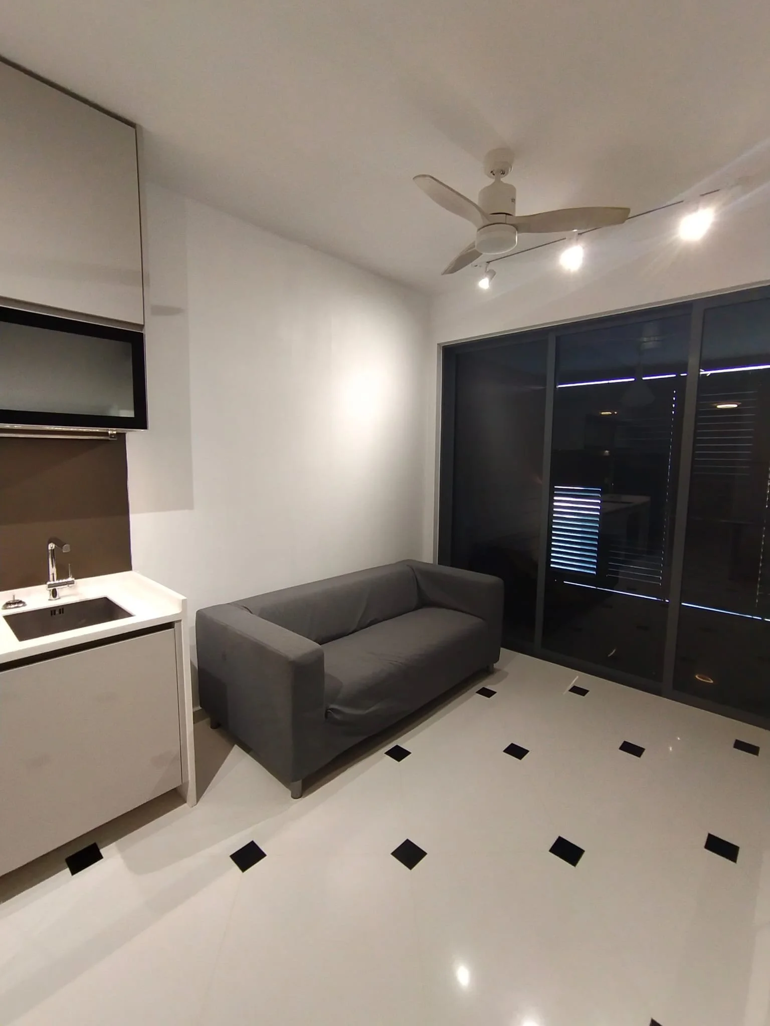 Minimalist living room with a gray sofa, black and white tiled flooring, a small kitchenette with a sink, a large sliding glass door leading outside, a ceiling fan, and ceiling lights.