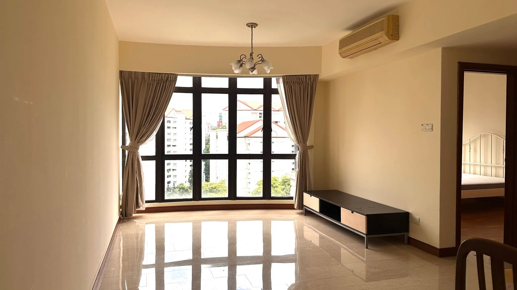 Empty living room with large window, beige walls, ceiling light fixture, and air conditioning unit. There's a black TV stand along the right wall and a doorway leading to a bedroom with a white bed frame.