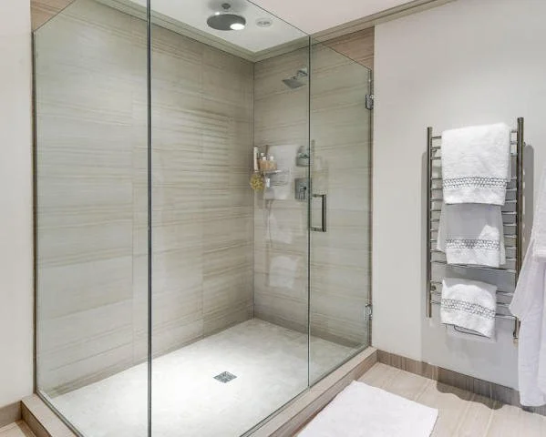 Empty walk-in shower with beige tiled walls and a glass door, white towel on a heated towel rack, and a white bath mat on the floor.