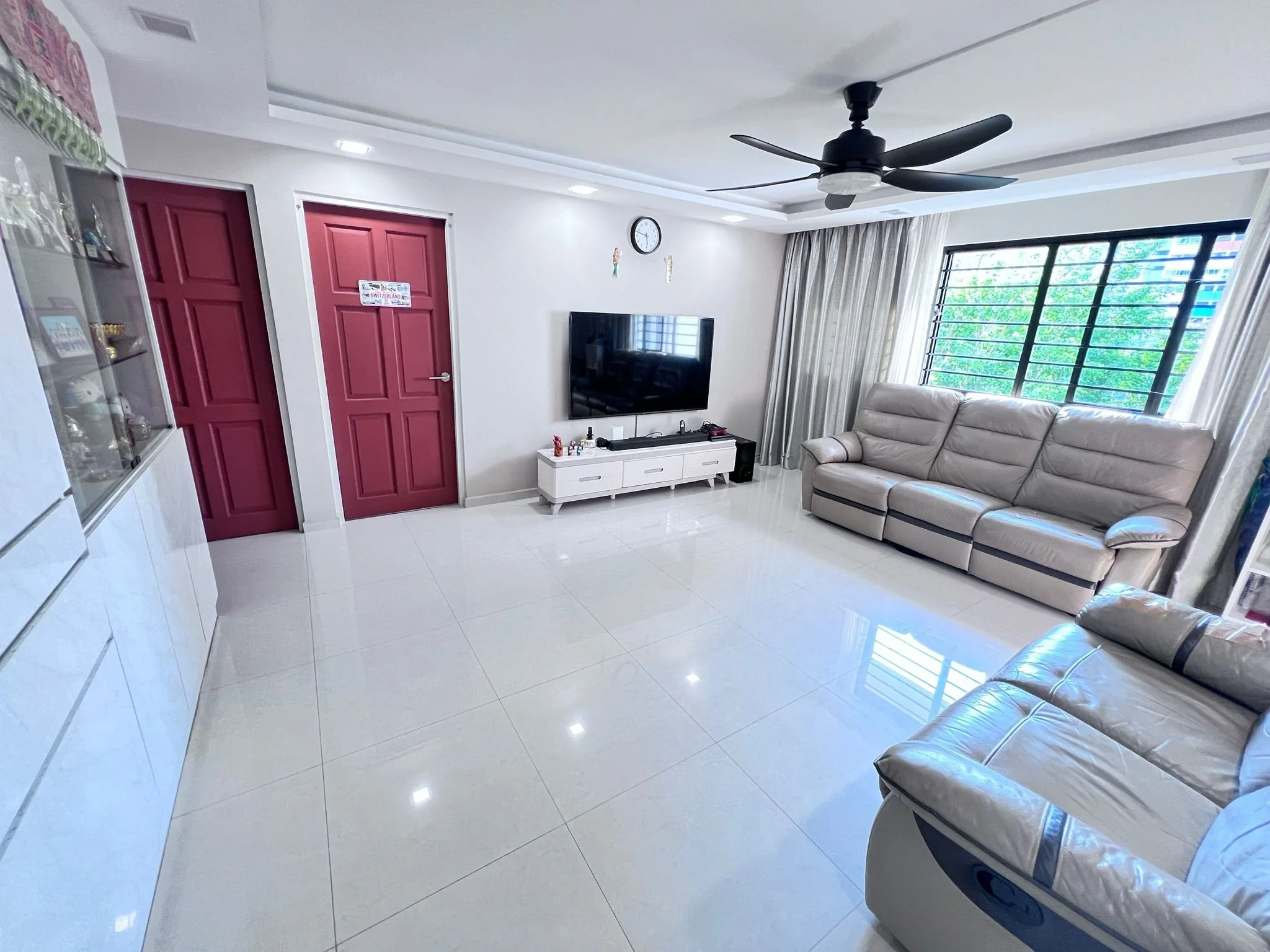 Bright living room with white tiled floors, beige sofas, a wall-mounted TV on a white stand, large window with white curtains, and red doors.