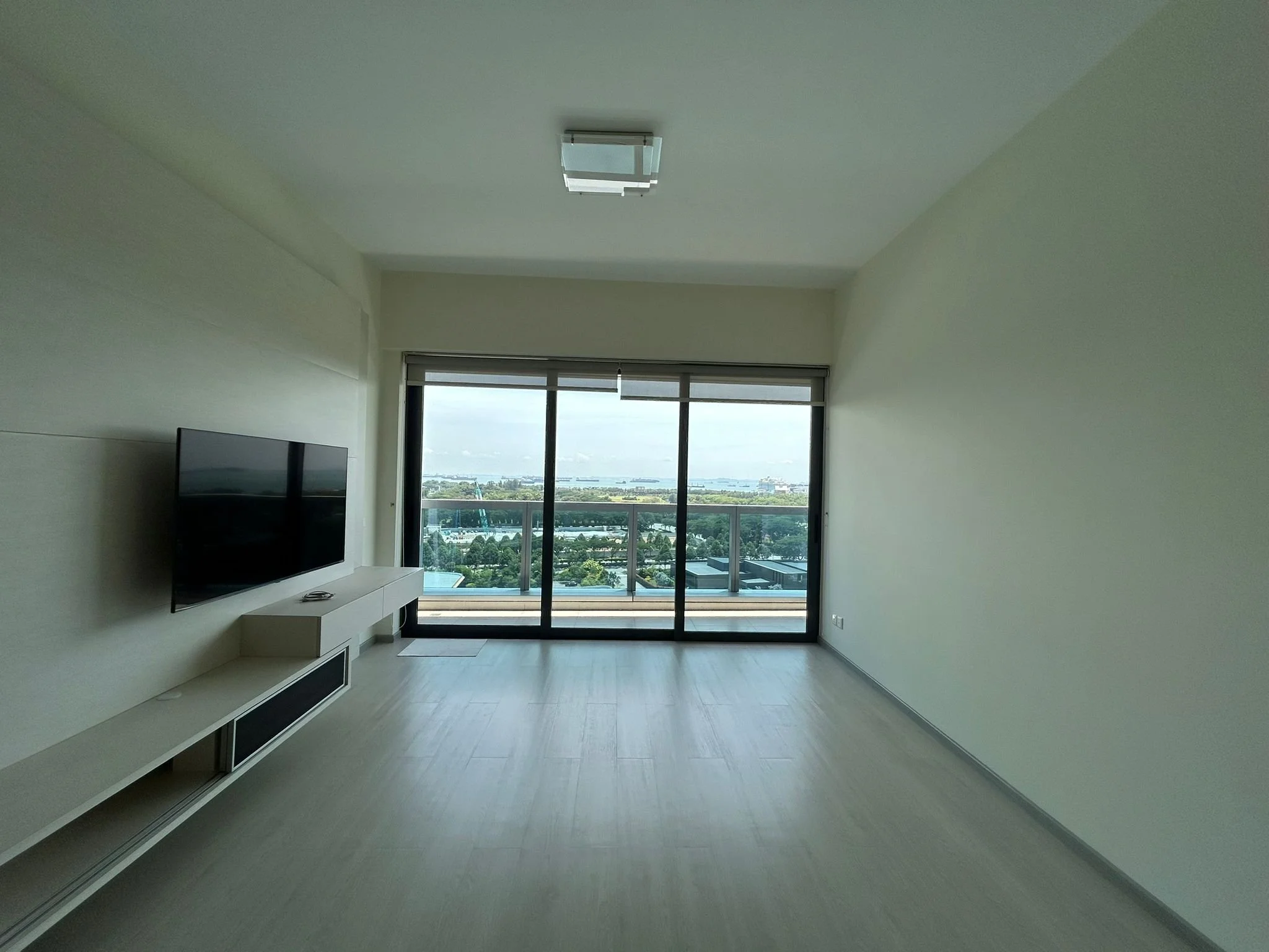 Empty living room with large sliding glass door overlooking city and water, minimal white walls and flooring, wall-mounted flat-screen TV.