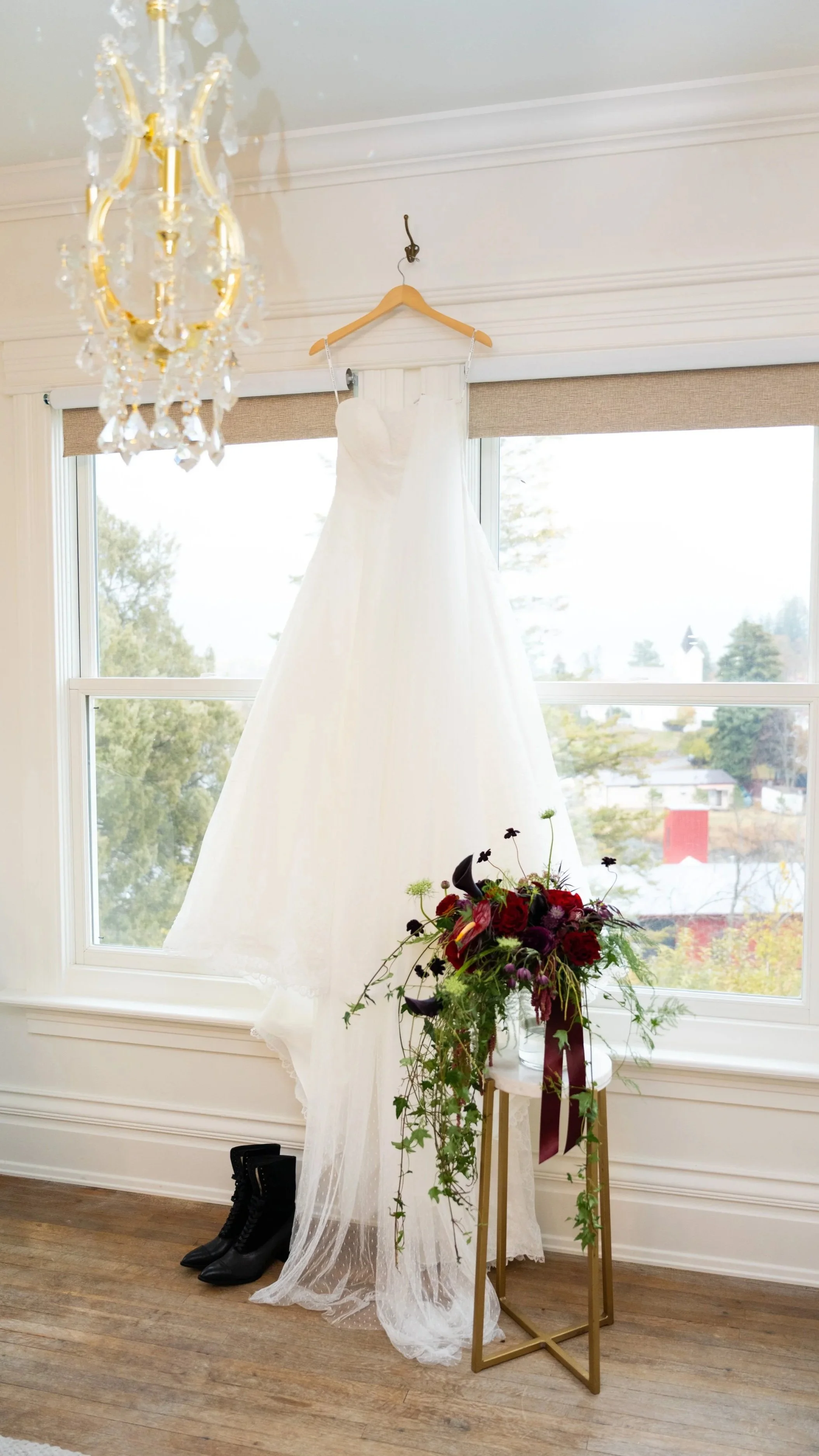 A white wedding dress hanging on a wooden hanger from a wall hook, with black ankle boots on the floor beneath it, in front of large windows. A gold and glass chandelier hangs from the ceiling, and a potted plant with red and purple flowers and greenery on a gold stand is nearby.