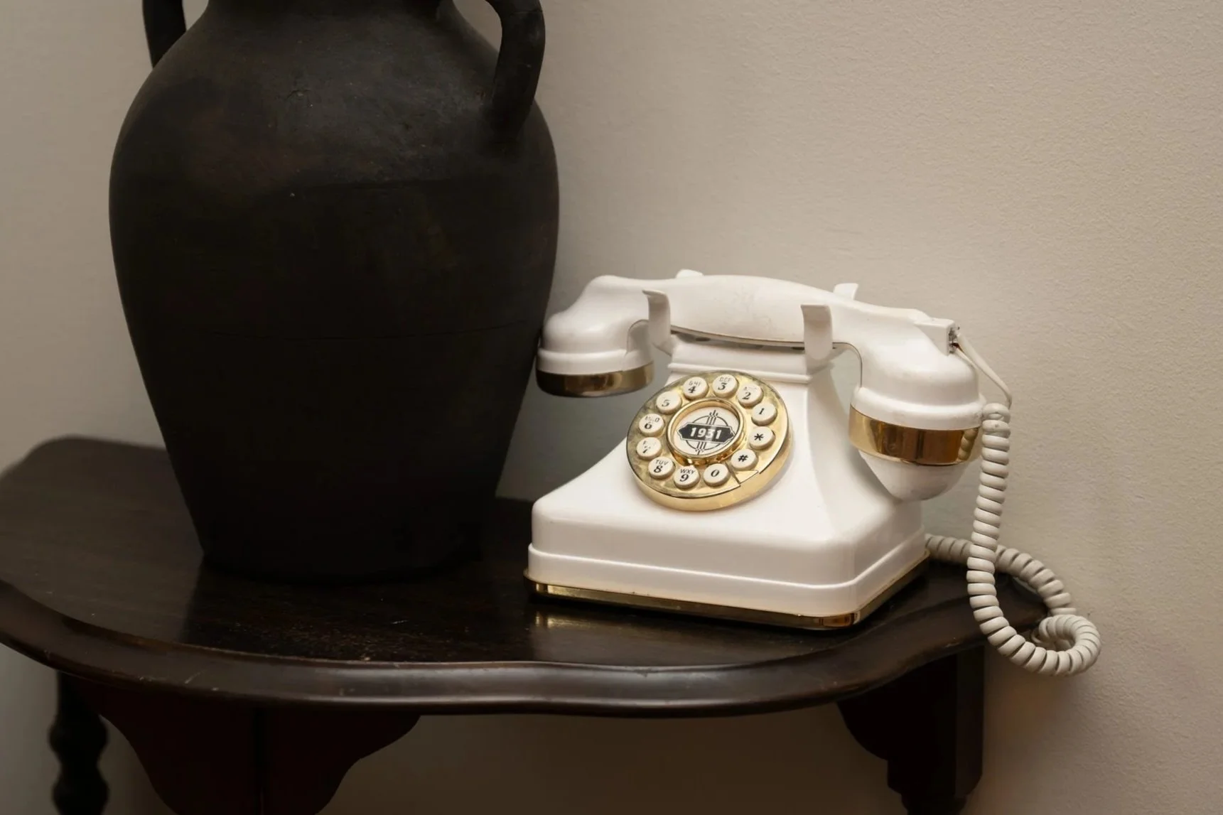A white vintage rotary telephone with gold accents on a dark wooden table, next to a large black vase against a plain wall.