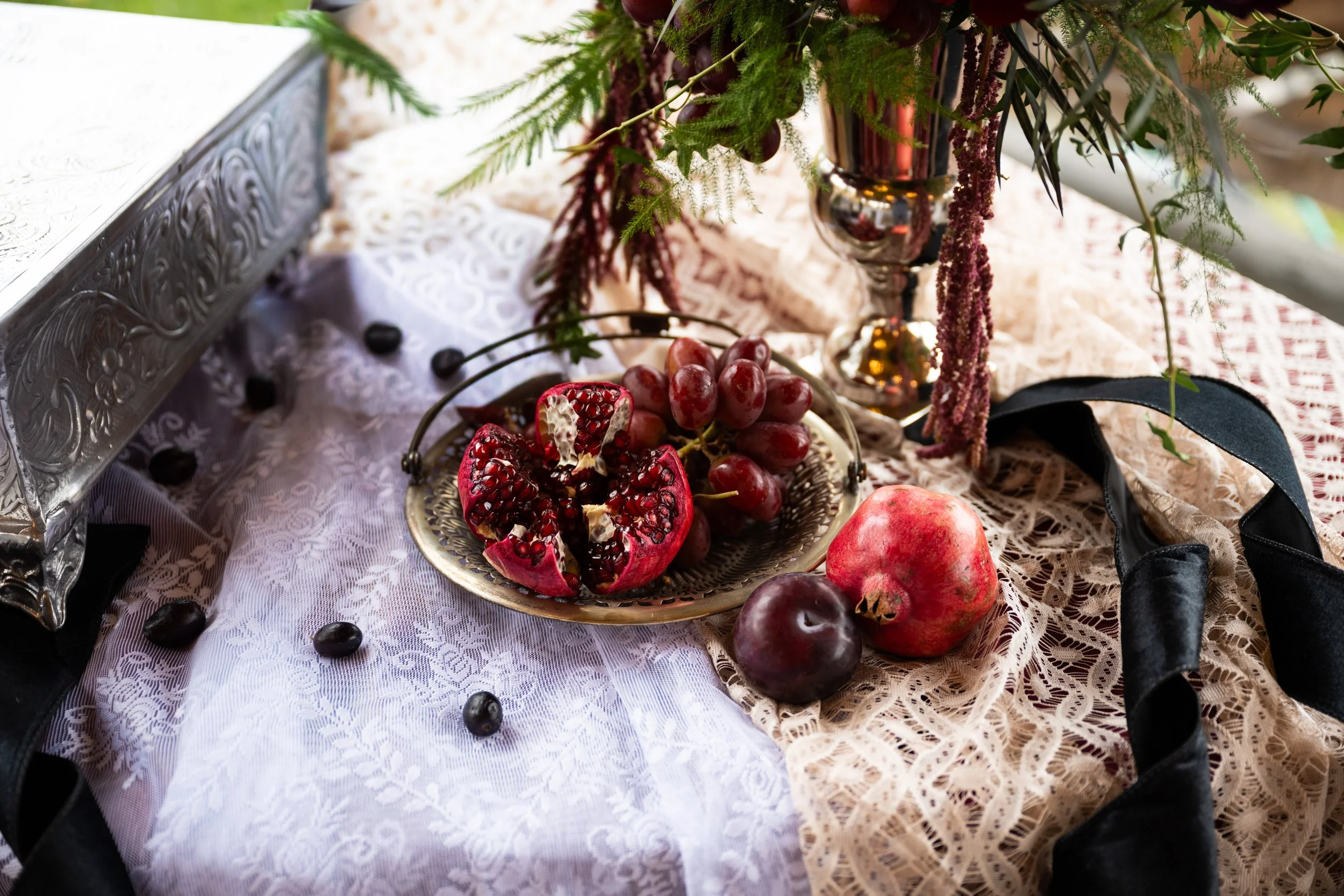 A decorative table setting with a lace tablecloth, featuring a silver tray with fresh pomegranate and grapes, a whole pomegranate and plum nearby, and a floral arrangement in a vase.