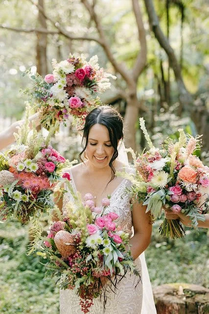 Bride holding multiple large, colorful floral bouquets outdoors in a wooded area.