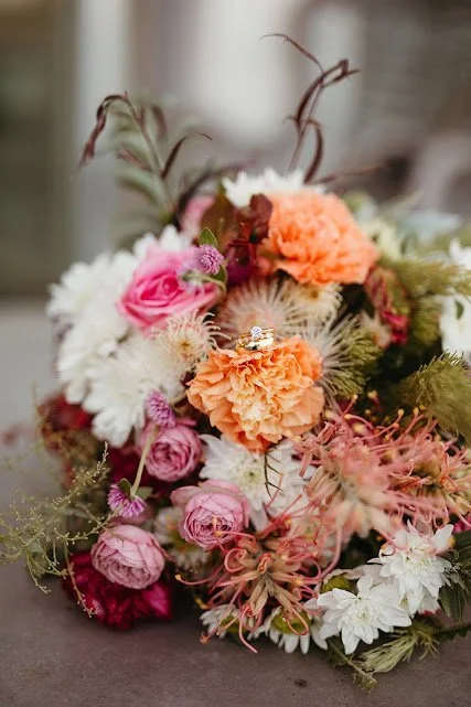 A bouquet of mixed flowers including pink roses, orange carnations, white flowers, and greenery.