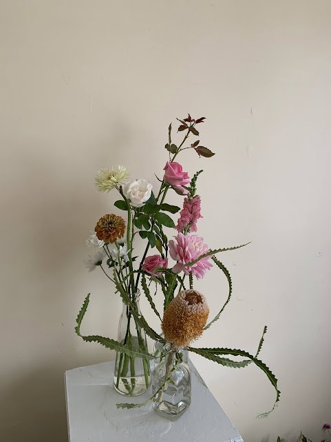 A glass vase with various flowers, including pink, white, and yellow blooms, placed on a white surface against a plain wall.
