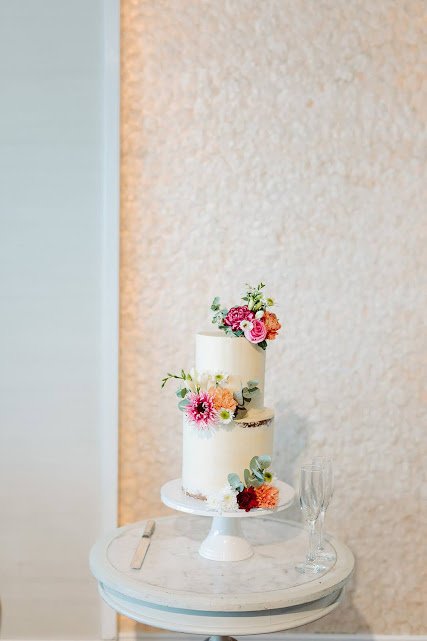 A white tiered wedding cake decorated with pink, orange, and peach flowers, placed on a round table with a cake knife and a champagne flute.