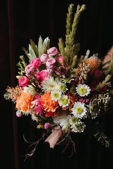 A colorful bouquet of various flowers, including pink roses, white daisies, orange carnations, and greenery, against a dark background.