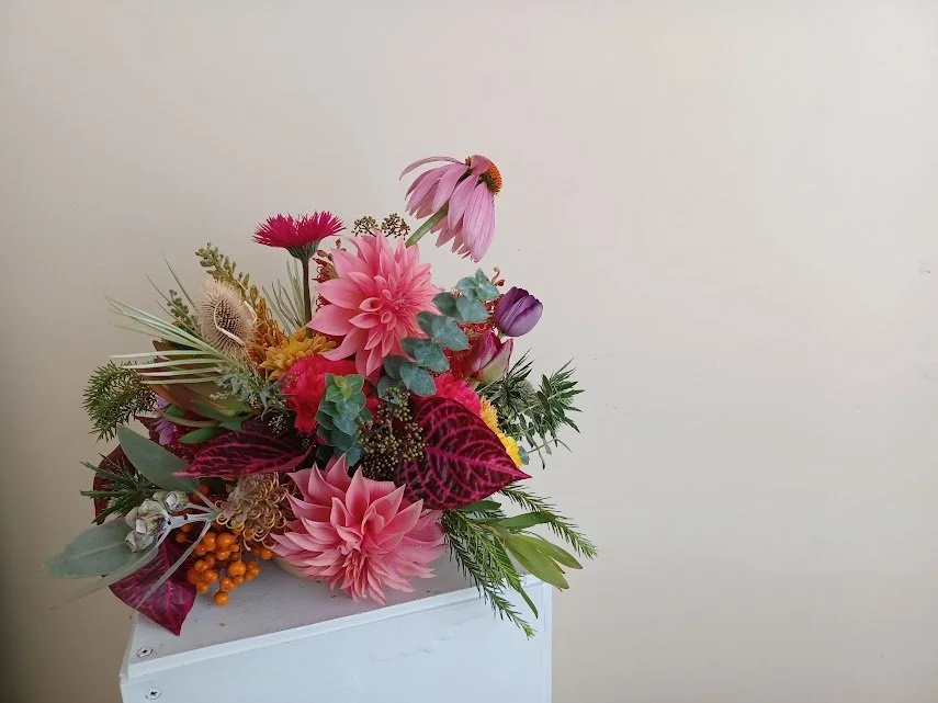Colorful floral arrangement with pink dahlias, coneflowers, eucalyptus, red and purple leaves, and other assorted flowers on a white surface against a plain beige wall.