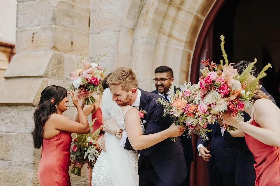 Small wedding party exchanging bouquets outside a stone building with an arched doorway.