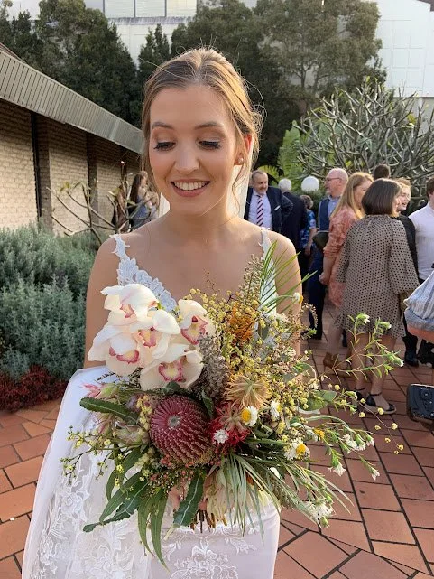 Woman in a white dress smiling and holding a large colorful bouquet of flowers outdoors at a gathering.
