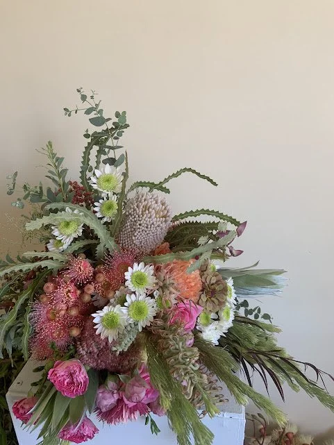 A large bouquet of mixed flowers including pink carnations, white daisies with green centers, and various greenery, arranged in a white box against a plain light-colored wall.