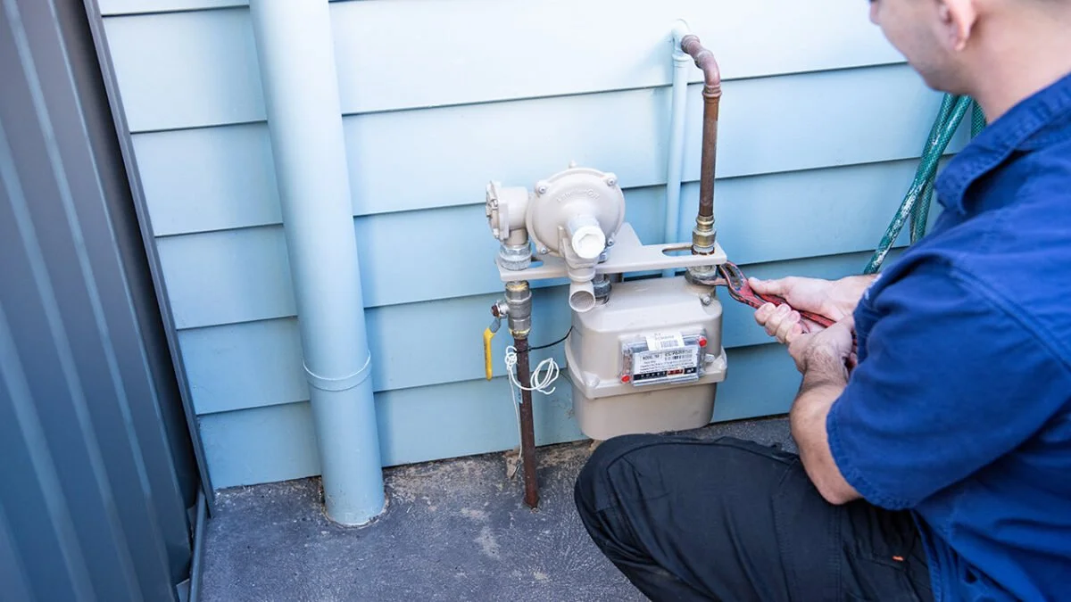 Technician working on a gas meter outside a house, holding a wrench with blue siding and a drain pipe visible.