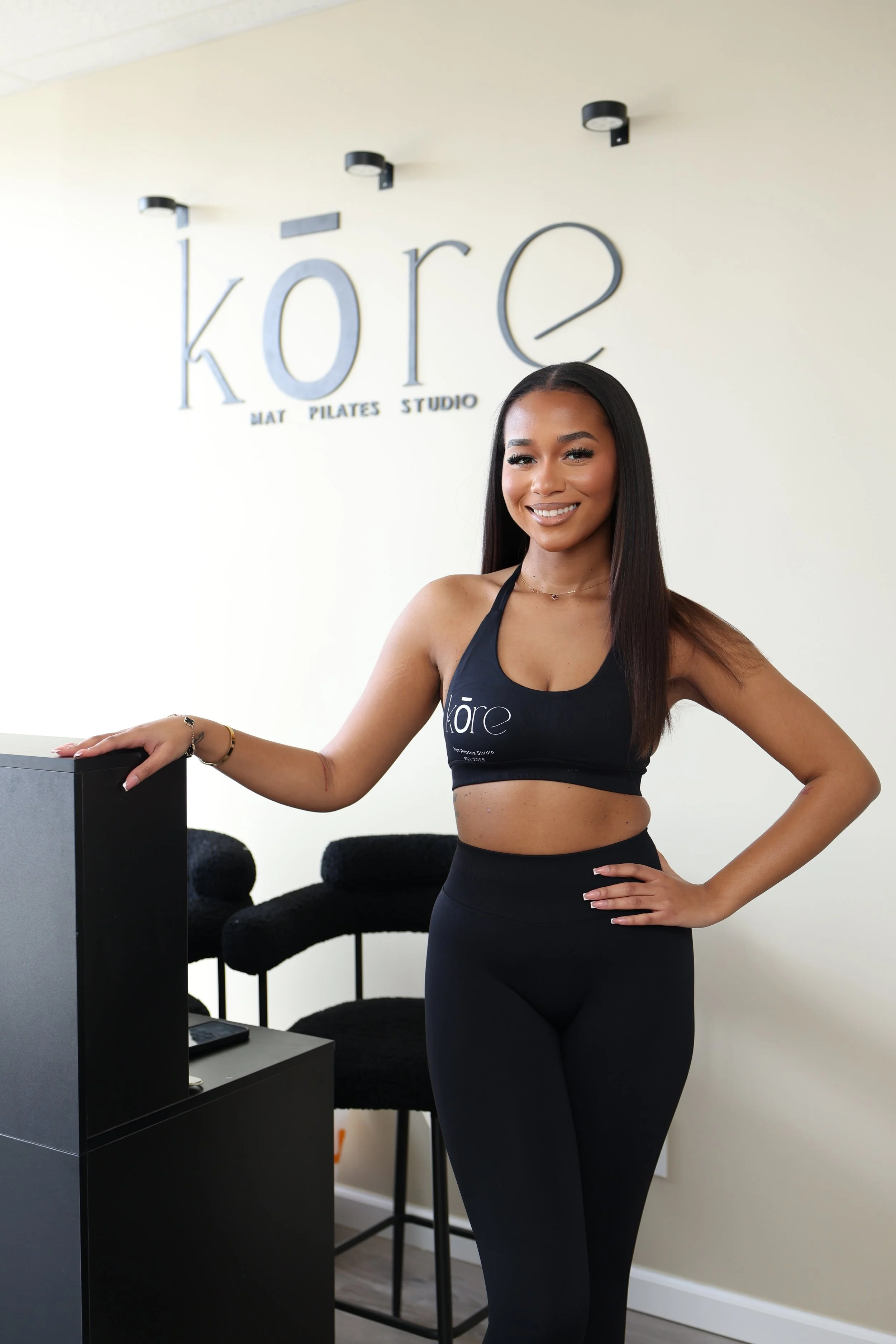 A woman wearing a black sports bra with the word 'kore' on it, smiling and standing in a fitness studio with black chairs behind her and a sign that reads 'kore MAT PILATES STUDIO' on the wall.