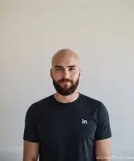 Man with a beard wearing a black t-shirt standing against a plain wall.