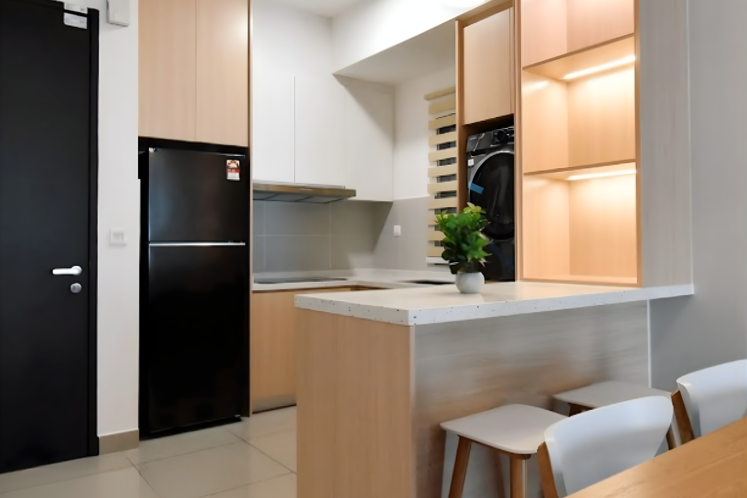 Modern kitchen with black refrigerator, wooden cabinets, and a small countertop with a potted plant. There are white chairs at a dining table visible in the foreground.