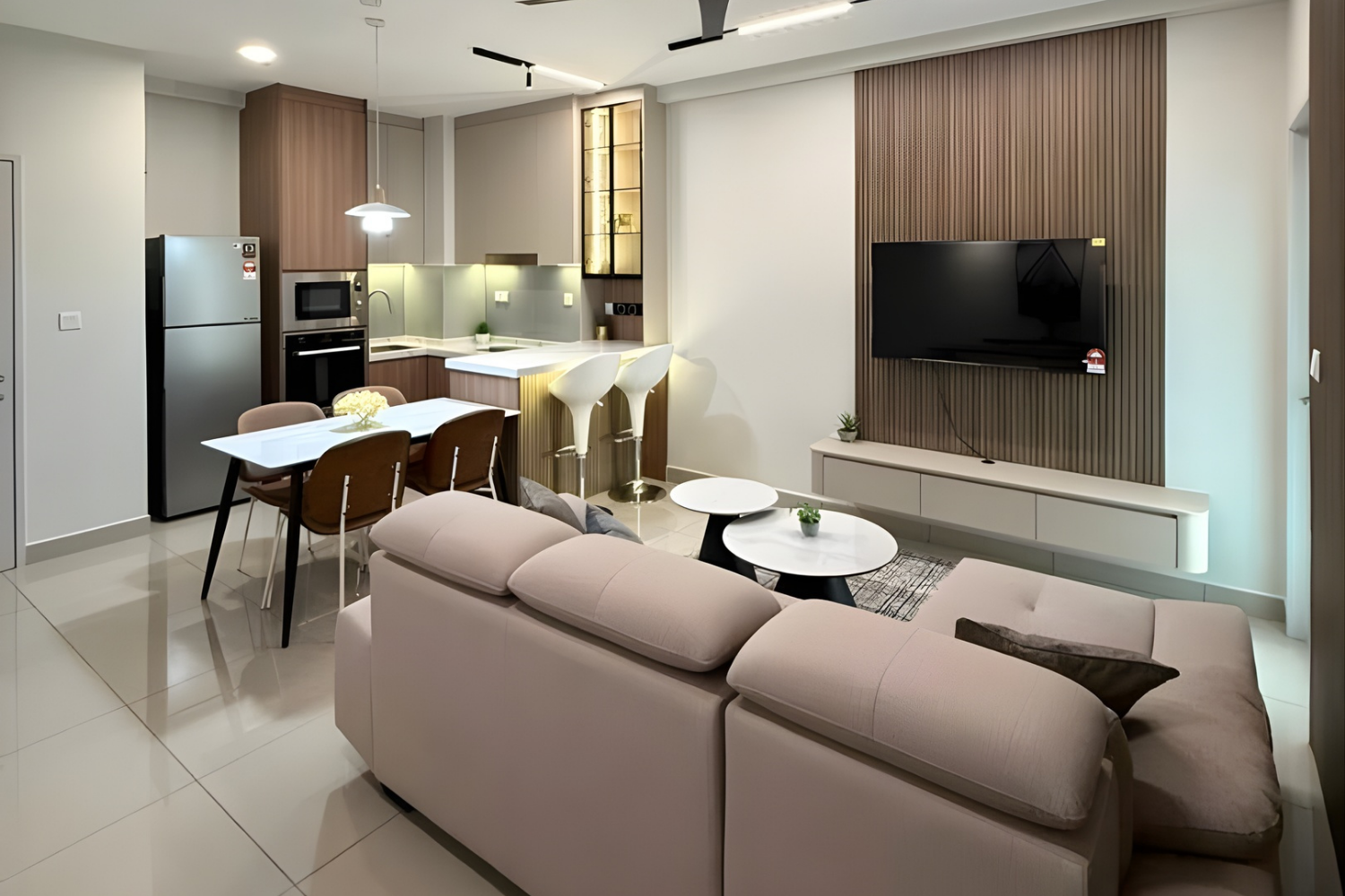 A modern open-concept living room and kitchen with white and wood accents, a beige sofa, a TV on a textured wood wall, a dining table with chairs, and a kitchen with appliances and a breakfast bar with two white barstools.
