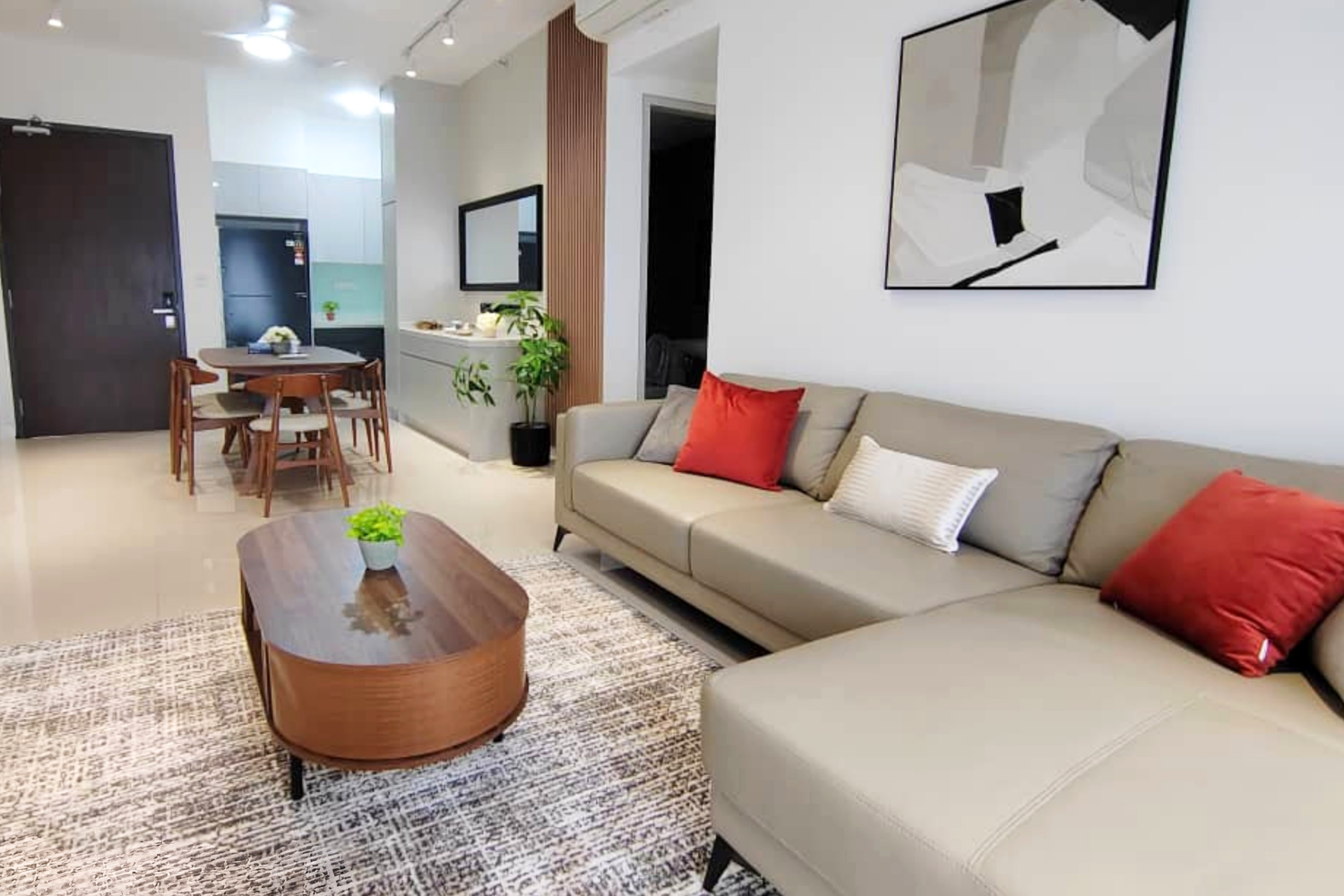 Living room with a beige sofa decorated with red and white pillows, a wooden coffee table with a small potted plant, and a patterned area rug. In the background, there's a dining area with a wooden table and chairs, and a small kitchen area with whit