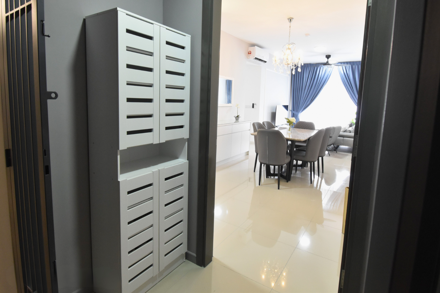 View into a modern dining and living room with a round dining table and six gray chairs, a white sideboard, a wall-mounted TV, and large blue curtains over a window.