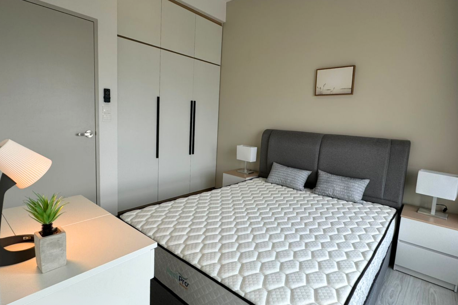 Modern bedroom with gray upholstered bed, two nightstands with white lamps, a white dresser with a potted plant and a desk lamp, a large closet with black handles, beige walls, and a simple framed piece of artwork.