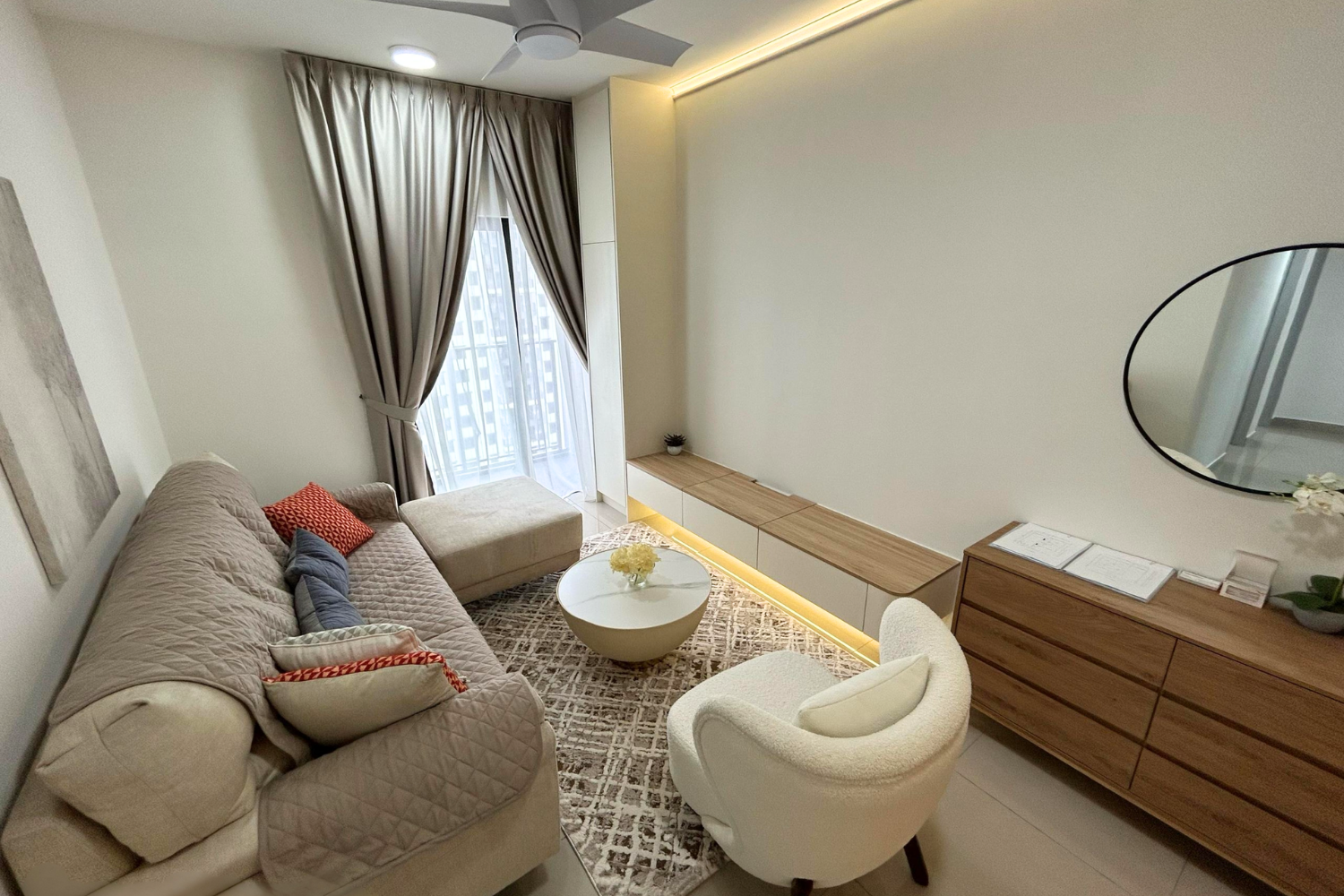 Living room with beige quilted sofa, white armchair, a round coffee table with flowers, large window with beige curtains, a long wooden TV console, wall-mounted mirror, and ceiling fan with lights.