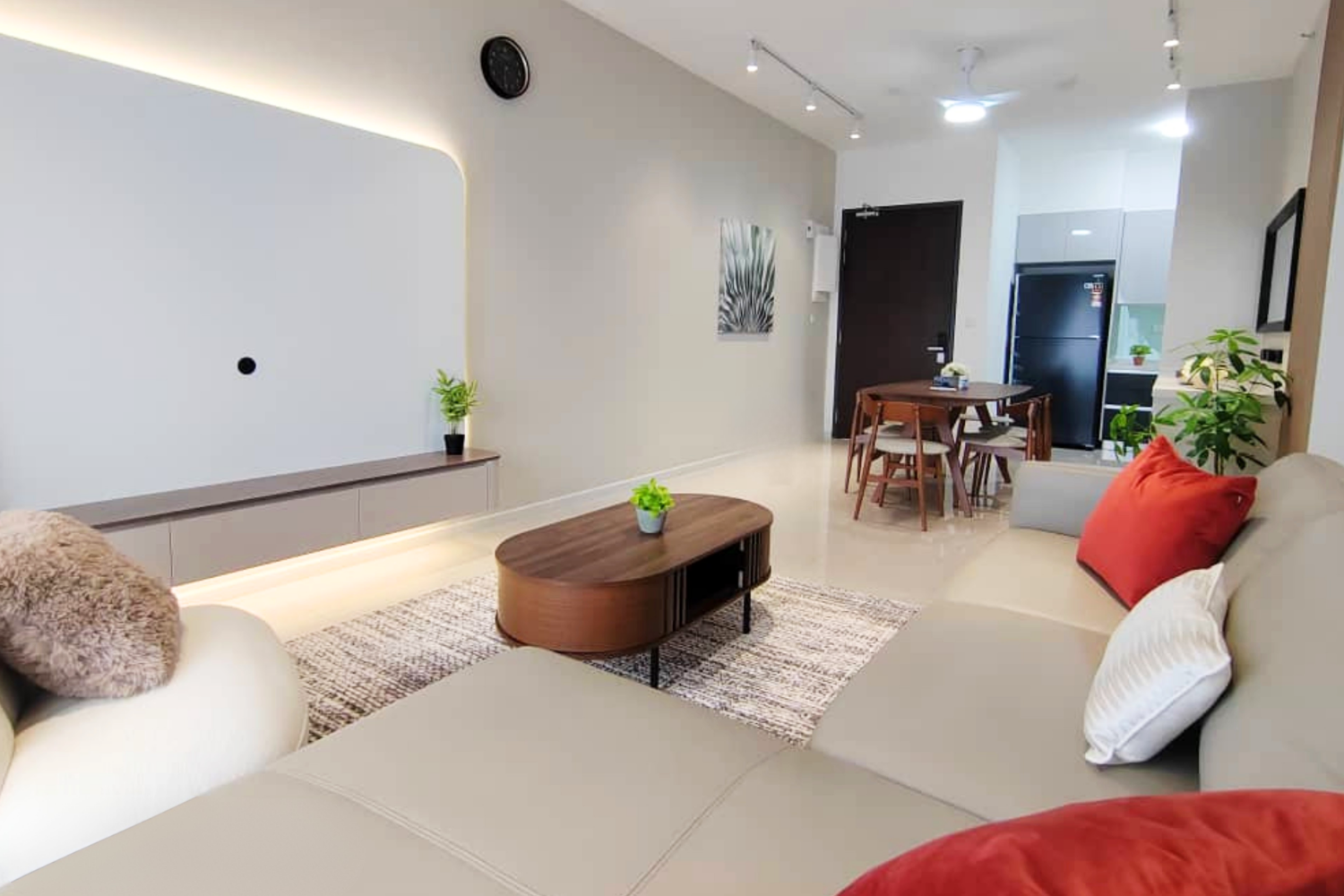 Living room with beige sofa and orange pillows, round wooden coffee table with a potted plant, white wall with wall clock, art piece, and TV, dining area with round table and chairs, black refrigerator, and kitchen with small potted plant.