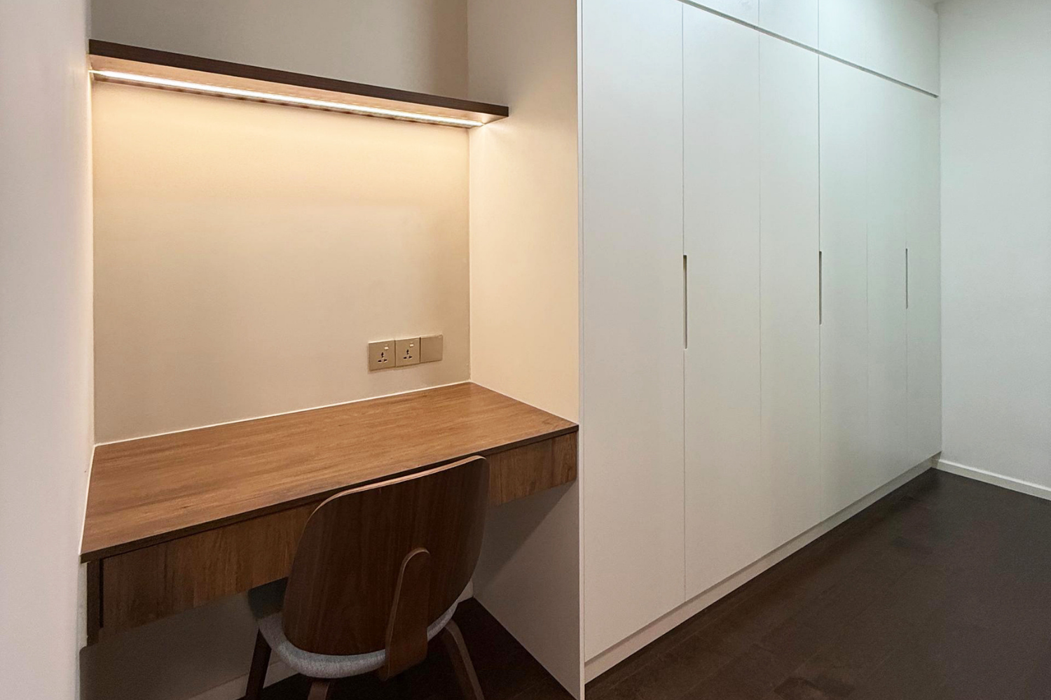 A built-in wooden desk with an office chair, a wall outlet, and shelving above it, adjacent to a large white wardrobe with multiple doors in a modern room.