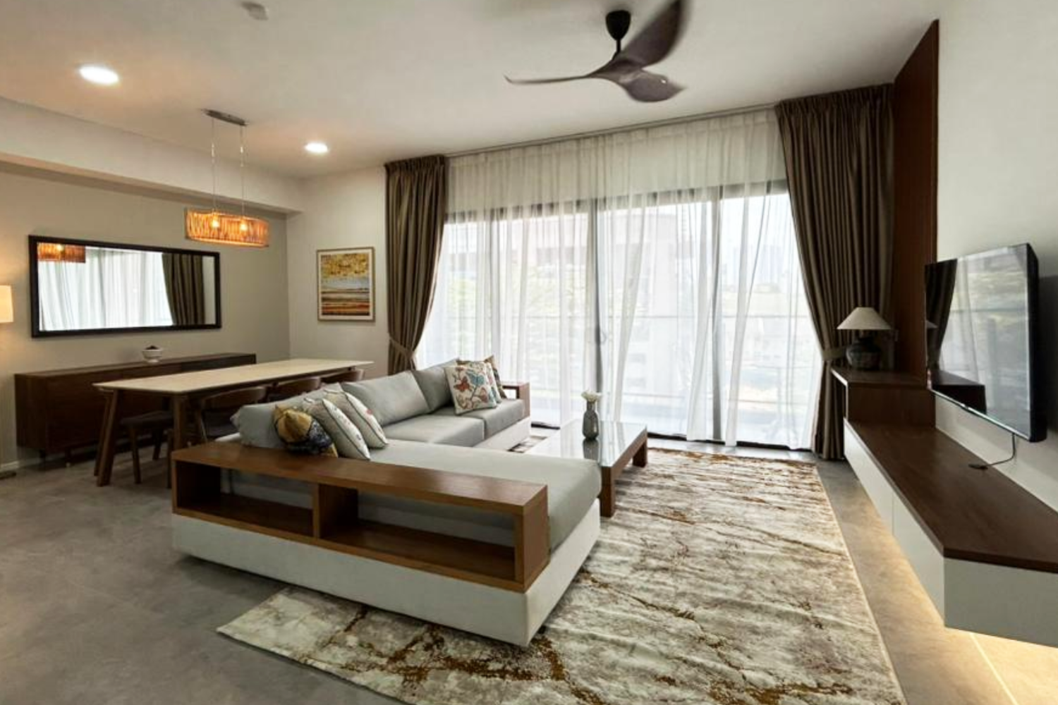 Modern living room with a light-colored sofa, decorative pillows, a wooden coffee table, large windows with sheer curtains, a flat-screen TV on a wooden shelf, and a dining area with a mirror and a wooden dining table.