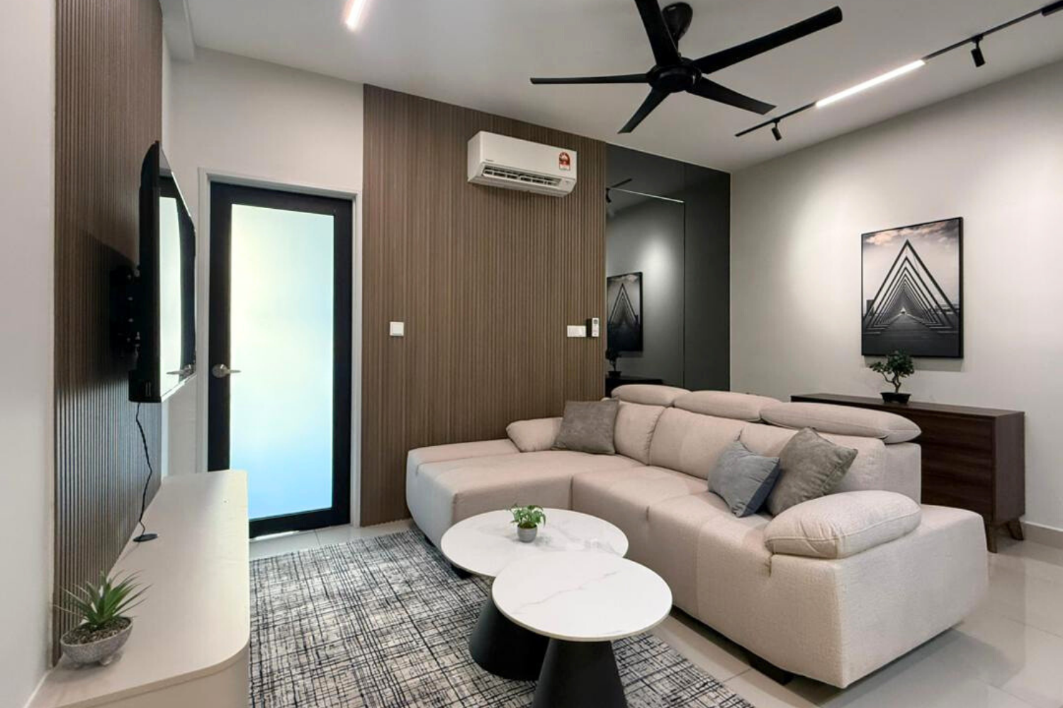 Modern living room with white sectional sofa, two round white and black coffee tables, black ceiling fan, wall-mounted TV, framed black-and-white artwork, small potted plant, air conditioner, and door with frosted glass.