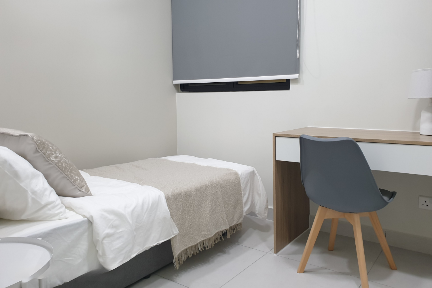 A bedroom with a bed, pillows, blankets, a window with a gray blind, a desk, and a blue chair.