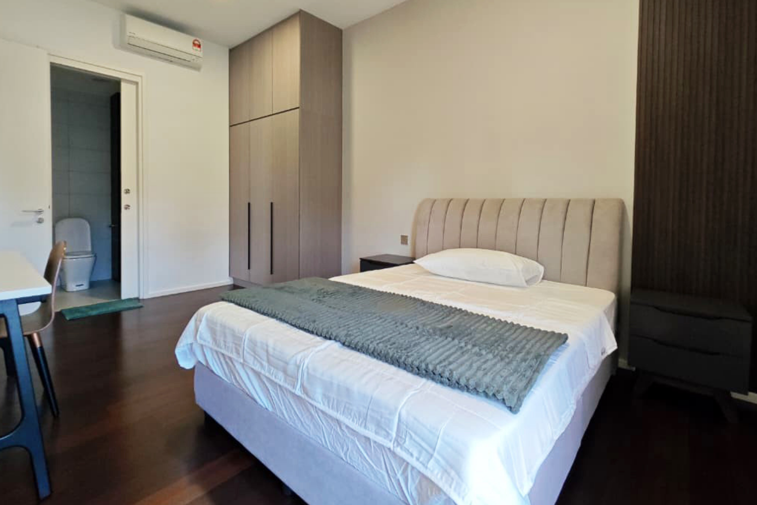 A bedroom with a bed, a beige upholstered headboard, a dark wooden nightstand, a large wardrobe, and a bathroom with a toilet visible through the doorway. The room has wooden flooring and neutral-colored walls.