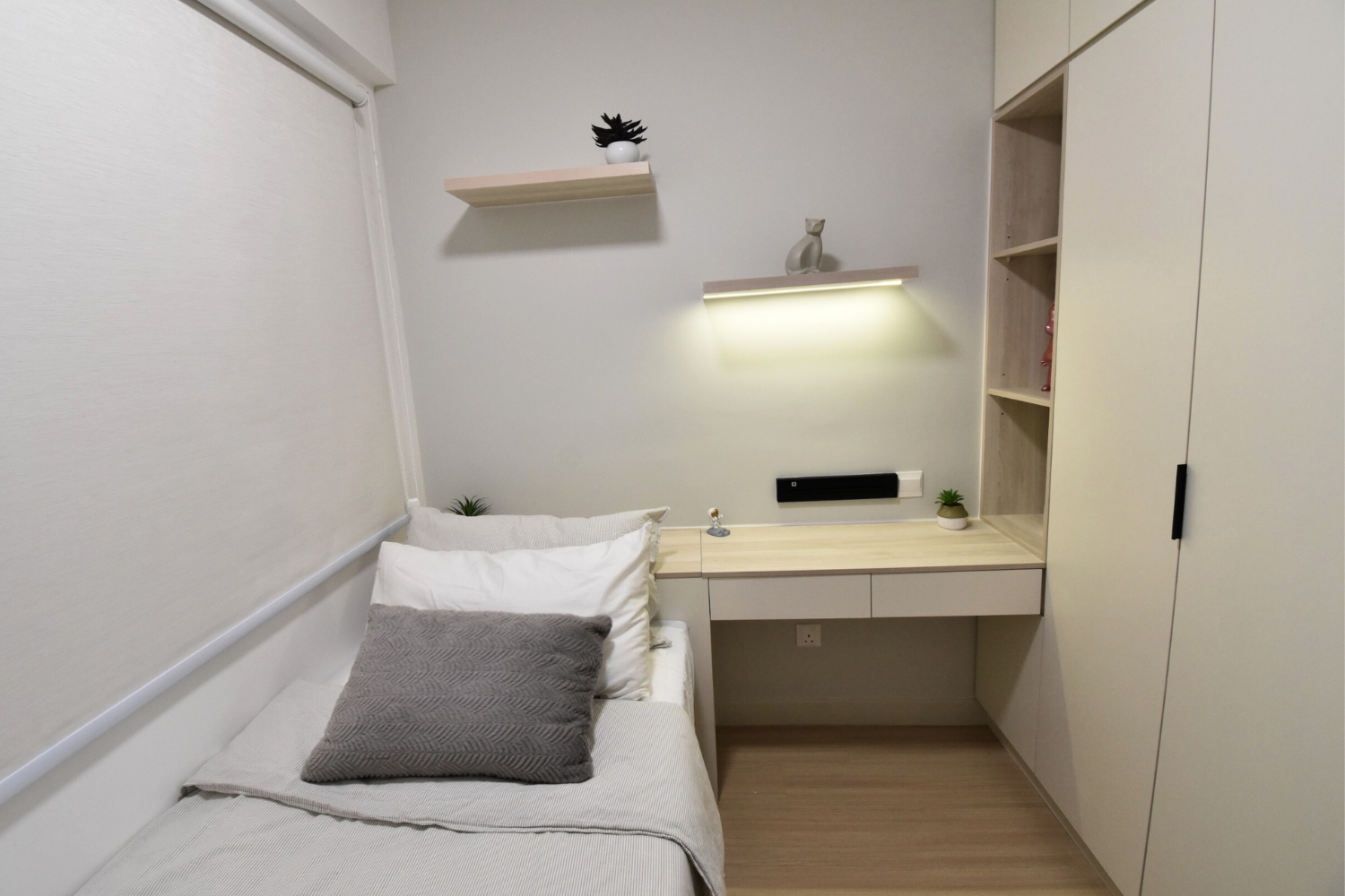 A minimalist bedroom with a single bed, gray pillow, white and beige bedding, a floating desk with small plants, decorative objects, and wall shelves with plants and a cat figurine.