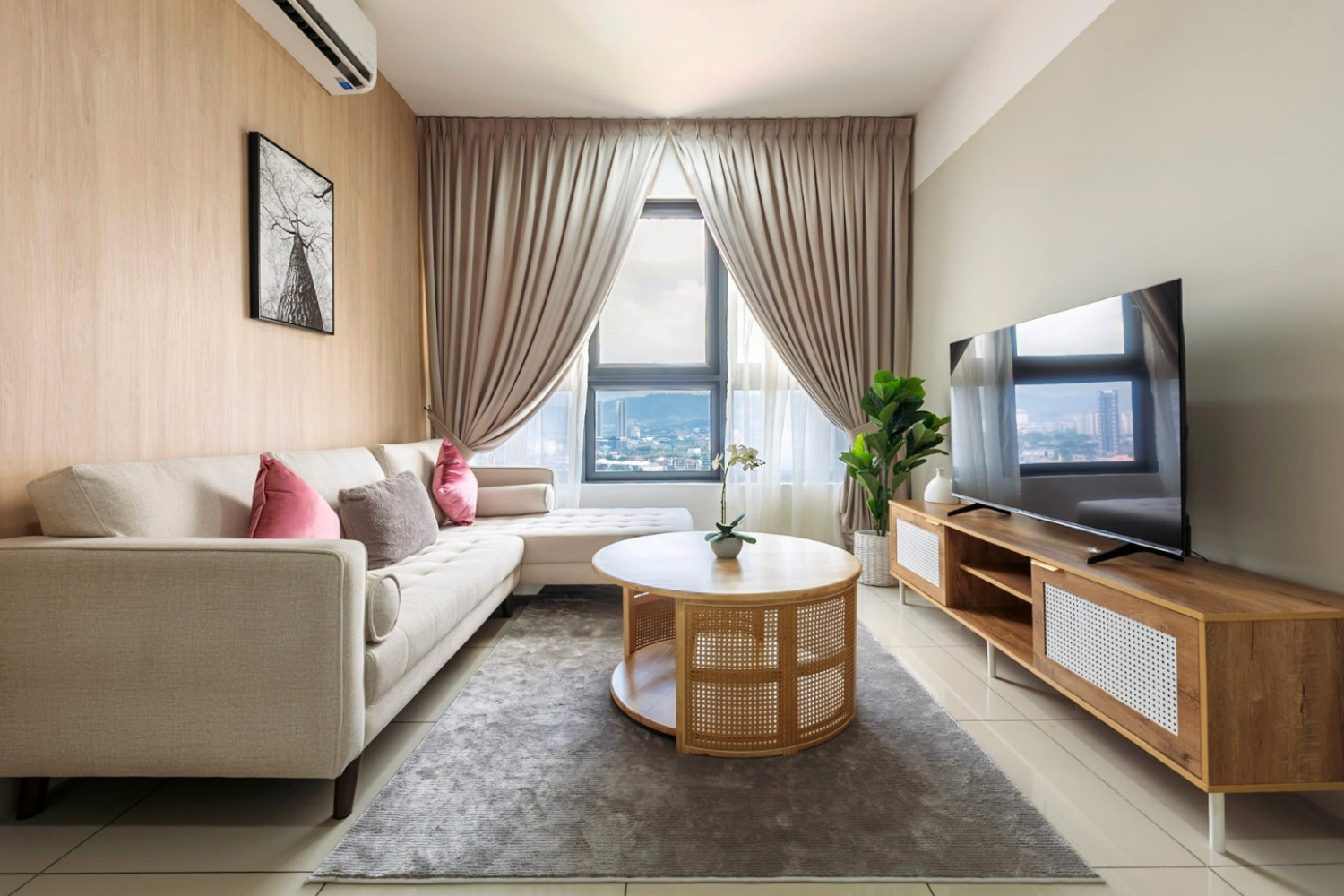 Modern living room with a beige sofa, pink and gray pillows, wooden TV stand, large window with curtains, coffee table with a plant, and a flat-screen TV.