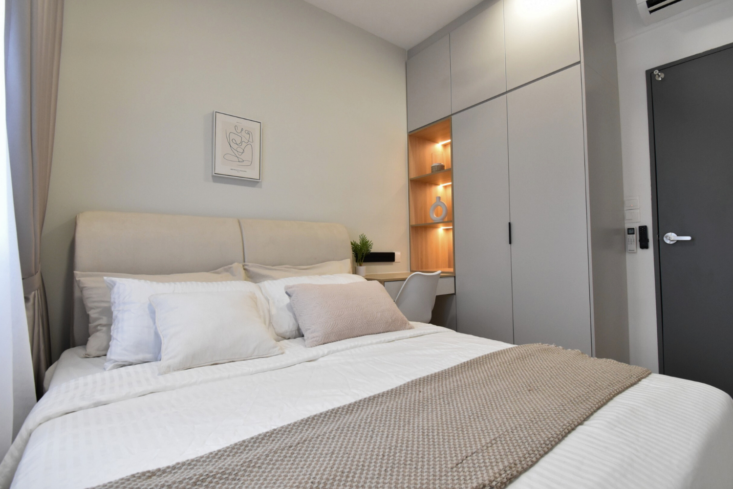 Modern bedroom with white bedding, a beige headboard, decorative pillows, a built-in wooden shelving unit with lighting, a potted plant, a desk, a white chair, gray closet doors, a dark gray door, and minimal decor.