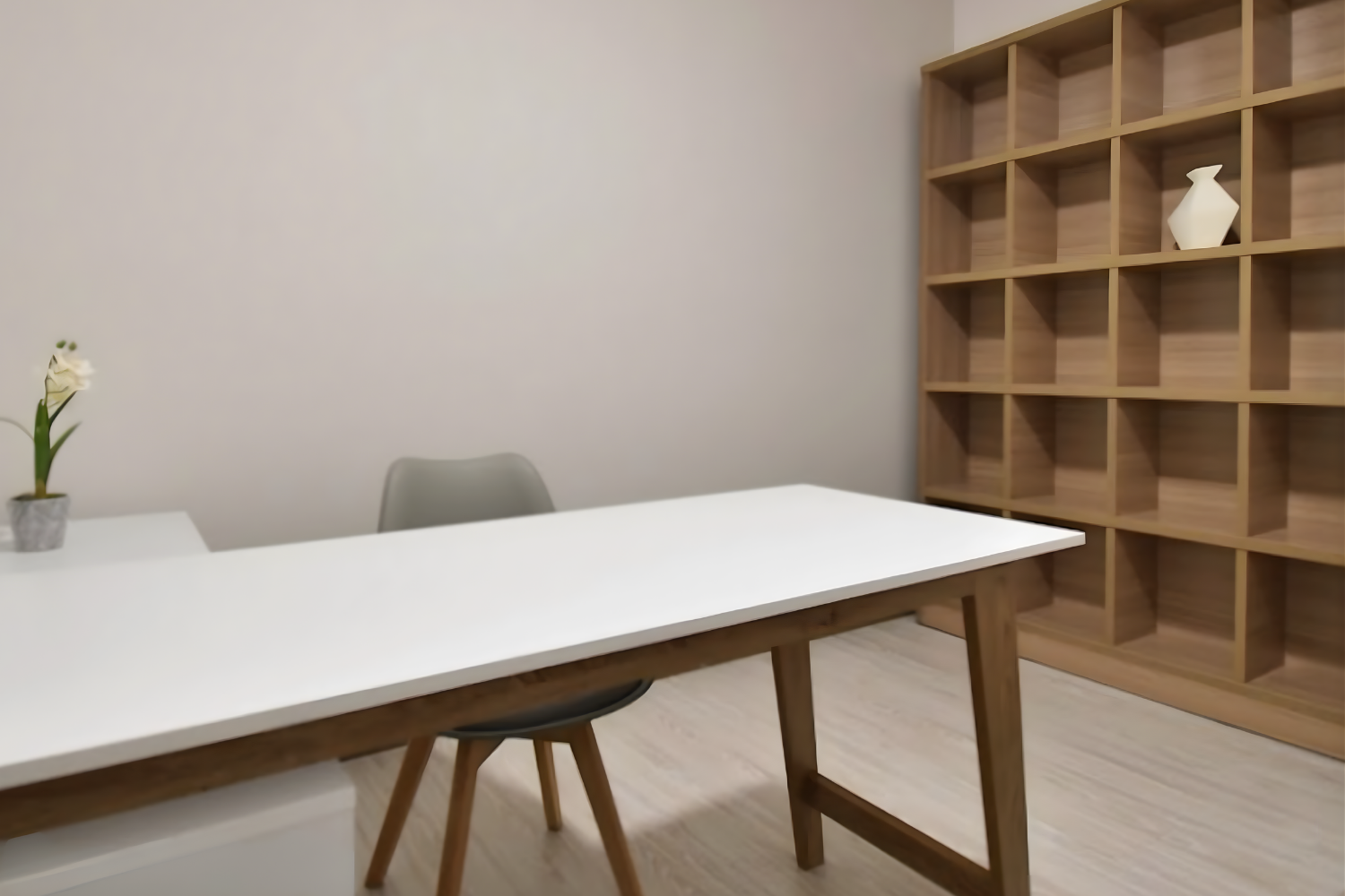 Minimalist office with white desk, gray chair, and a plant in a pot on the desk, beige wall, wooden shelving unit with one white vase, and light wood flooring.