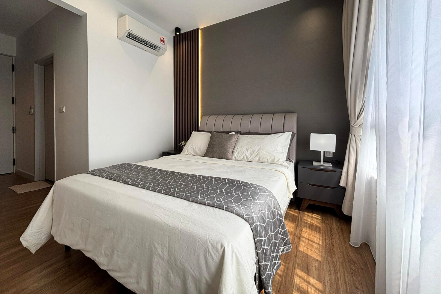 A modern bedroom with a large bed covered in white bedding and gray accents, situated against a dark accent wall with vertical wood paneling and lighting, with a bedside table, a small lamp, and a window with white curtains on the right.