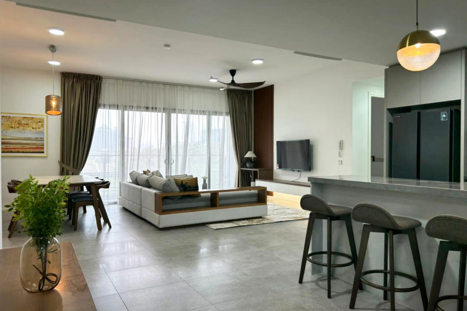 Modern living room with large window, white sofa, flat-screen TV, and dining area with plants.