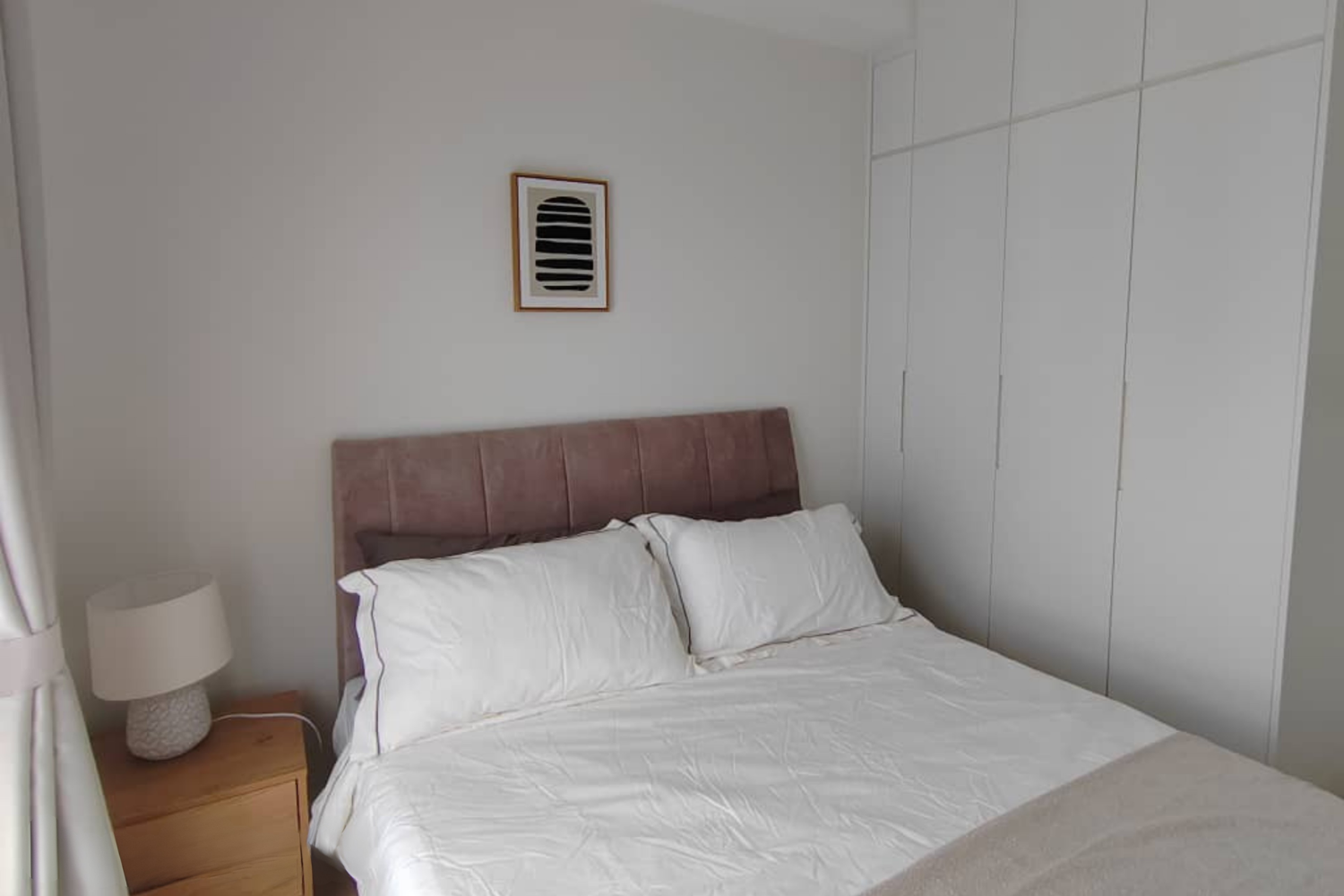 A bedroom with a bed, white bedding, and multiple pillows. There is a wooden nightstand with a white lamp on it, a framed abstract black and white art piece on the wall, and white built-in closets.