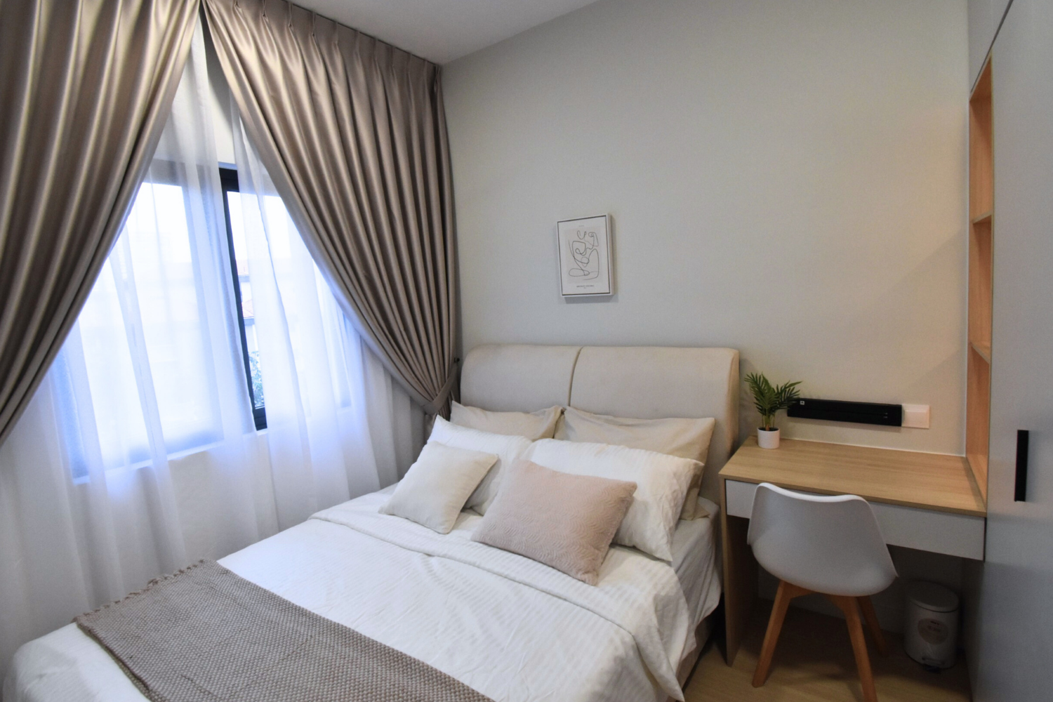 A cozy bedroom with a neatly made bed featuring white and beige pillows, a nightstand with a small plant, and a desk with a chair, beside a window with beige curtains and sheer white drapes.