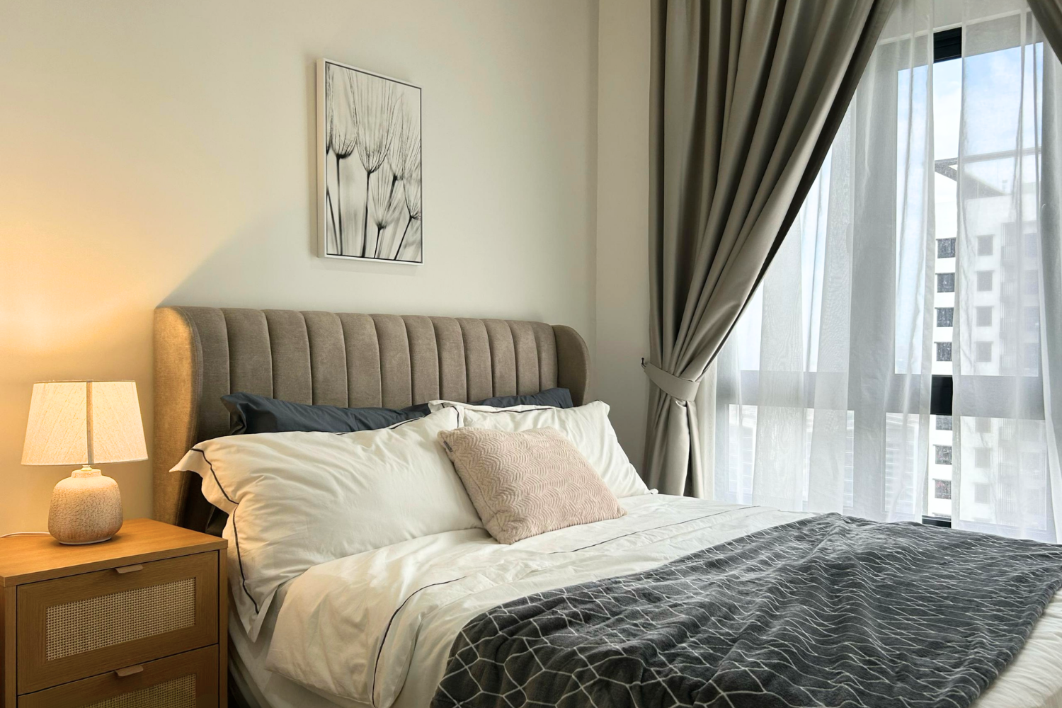 A neatly made bed with white linens, a beige tufted headboard, and decorative pillows, located in a bright bedroom with a window covered by sheer and draped curtains, and a nightstand with a table lamp beside the bed.