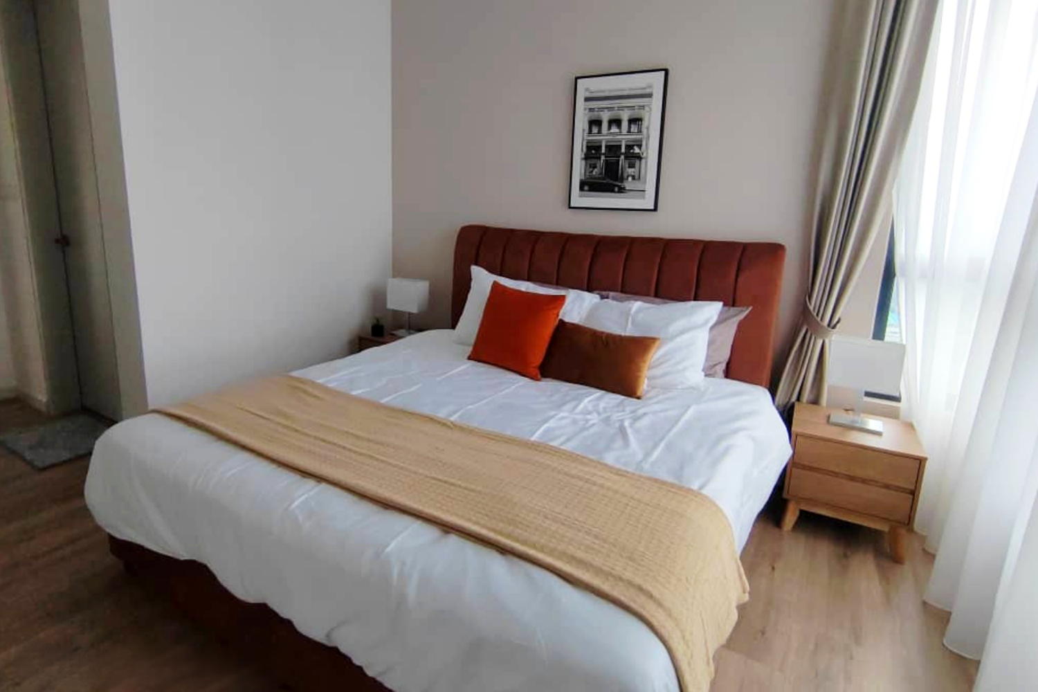 A neatly made bed with white bedding, a brown upholstered headboard, and three decorative pillows in burnt orange and caramel colors. Next to the bed is a wooden nightstand with a small lamp, and a framed black and white photograph hangs above the he
