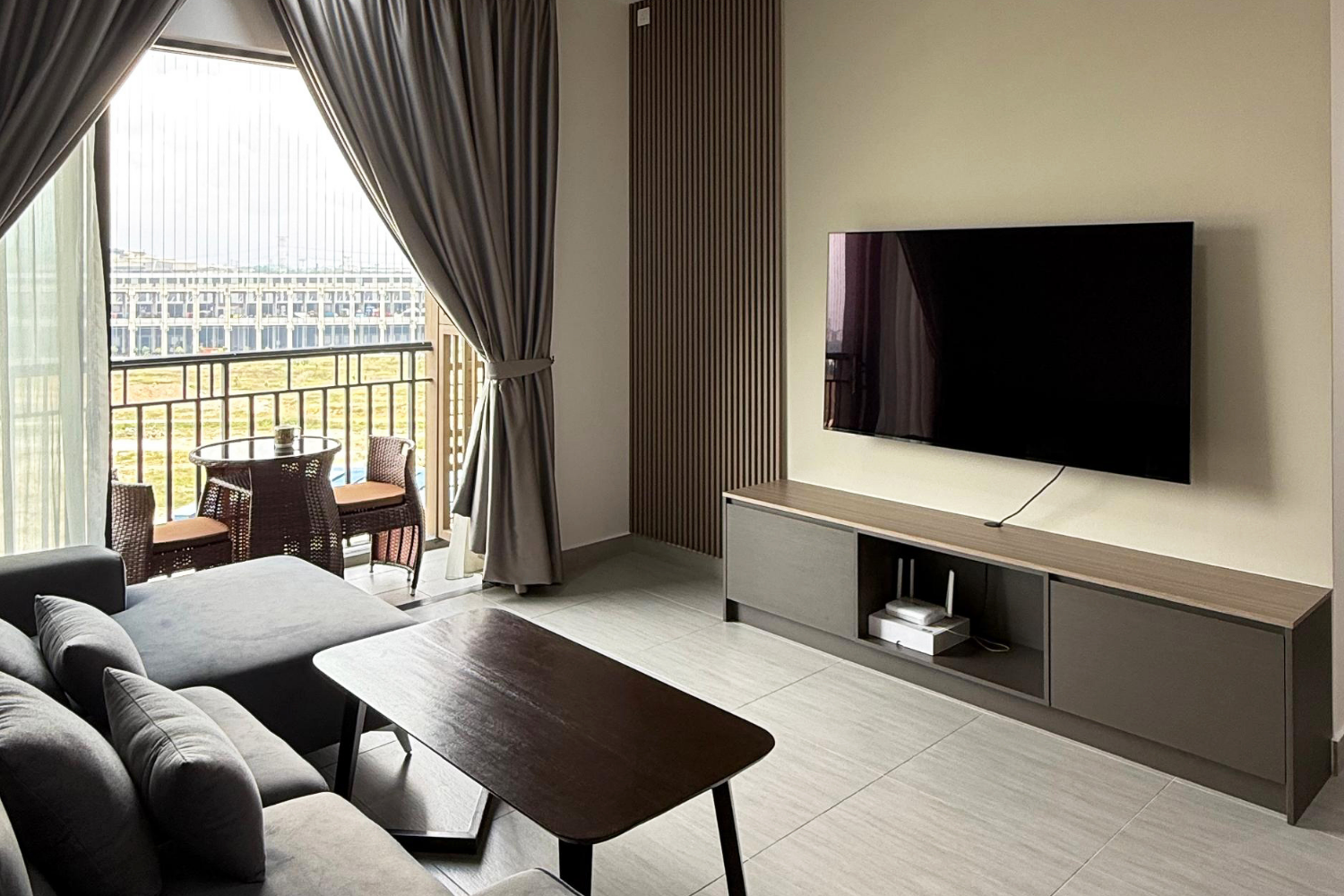 Living room with a brown coffee table, gray sofa, TV mounted on the wall, and a balcony with chairs and a table outside.