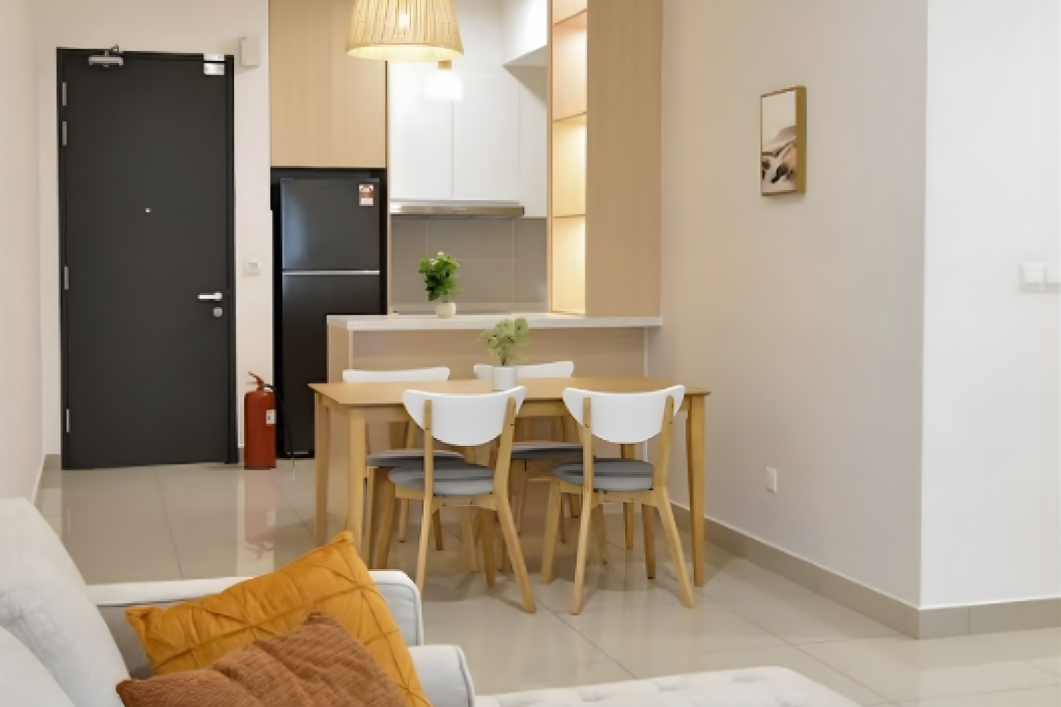 Modern living space with a dining area featuring a wooden table and six white chairs, a small kitchen with light wood cabinets and a black refrigerator, and a wall with a framed picture, illuminated by warm lighting.