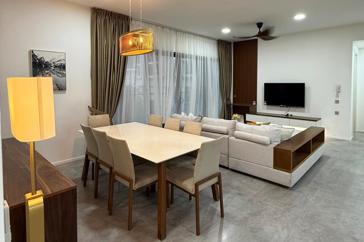 Modern living room with a dining table, beige chairs, a white sectional sofa, a wall-mounted flat-screen TV, large windows with curtains, a wooden ceiling fan, and contemporary decor.