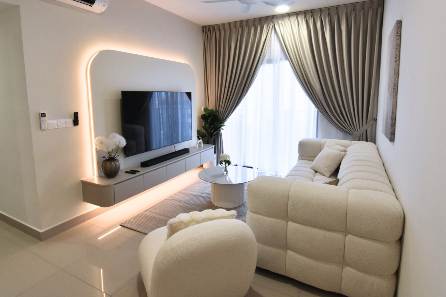 Modern living room with a large cream-colored sectional sofa, a matching armchair, and a round marble coffee table with flowers. There is a wall-mounted flat-screen TV with a soundbar below, a vase with white flowers, and a large window with beige st